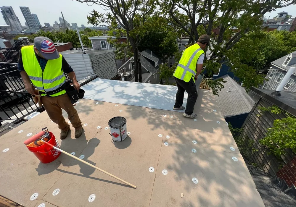 Two Alvarez Immediate Service roofers working on a white flat roof, with the Boston skyline behind them.