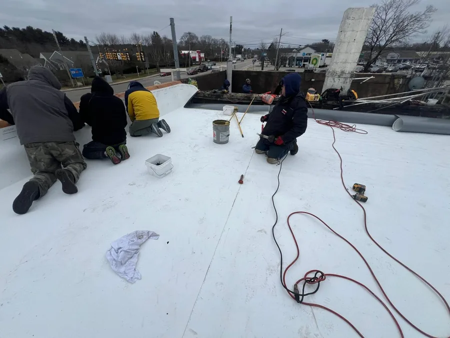 Four roofers kneeling on a large white-coated flat commercial roof, running hoses and coating equipment across the deck.