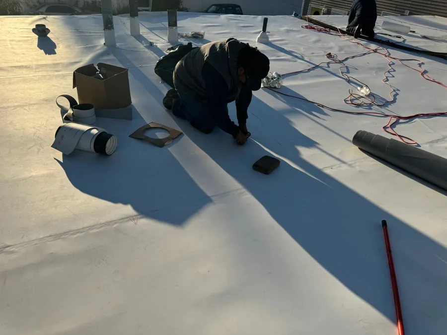 A roofer fitting single-ply membrane around vents and equipment curbs on a white flat roof.
