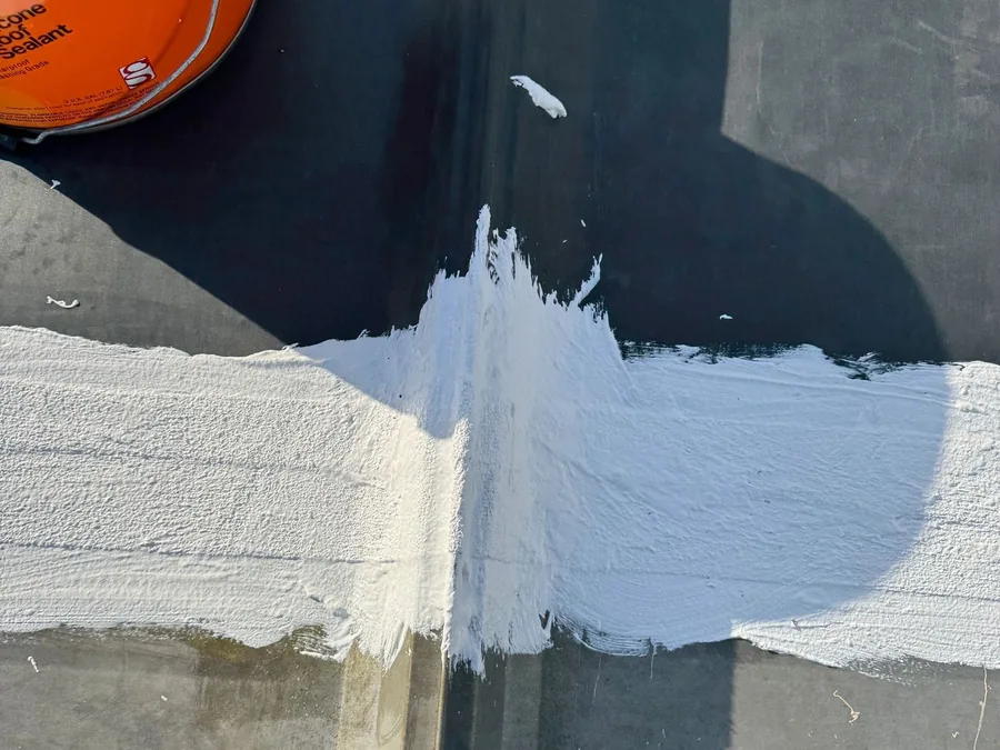 Close detail of a roof seam sealed and coated in white over the dark membrane below, with a coating tin alongside.